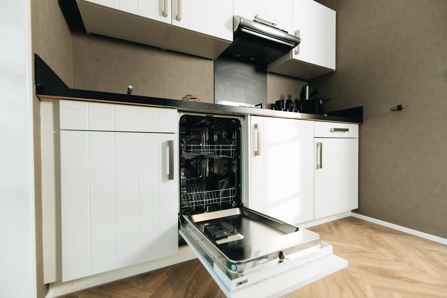 Detail van de keukenfaciliteiten in de lodge, met een geopende Zanussi vaatwasser, witte kastjes en een stijlvolle houtlook visgraatvloer.